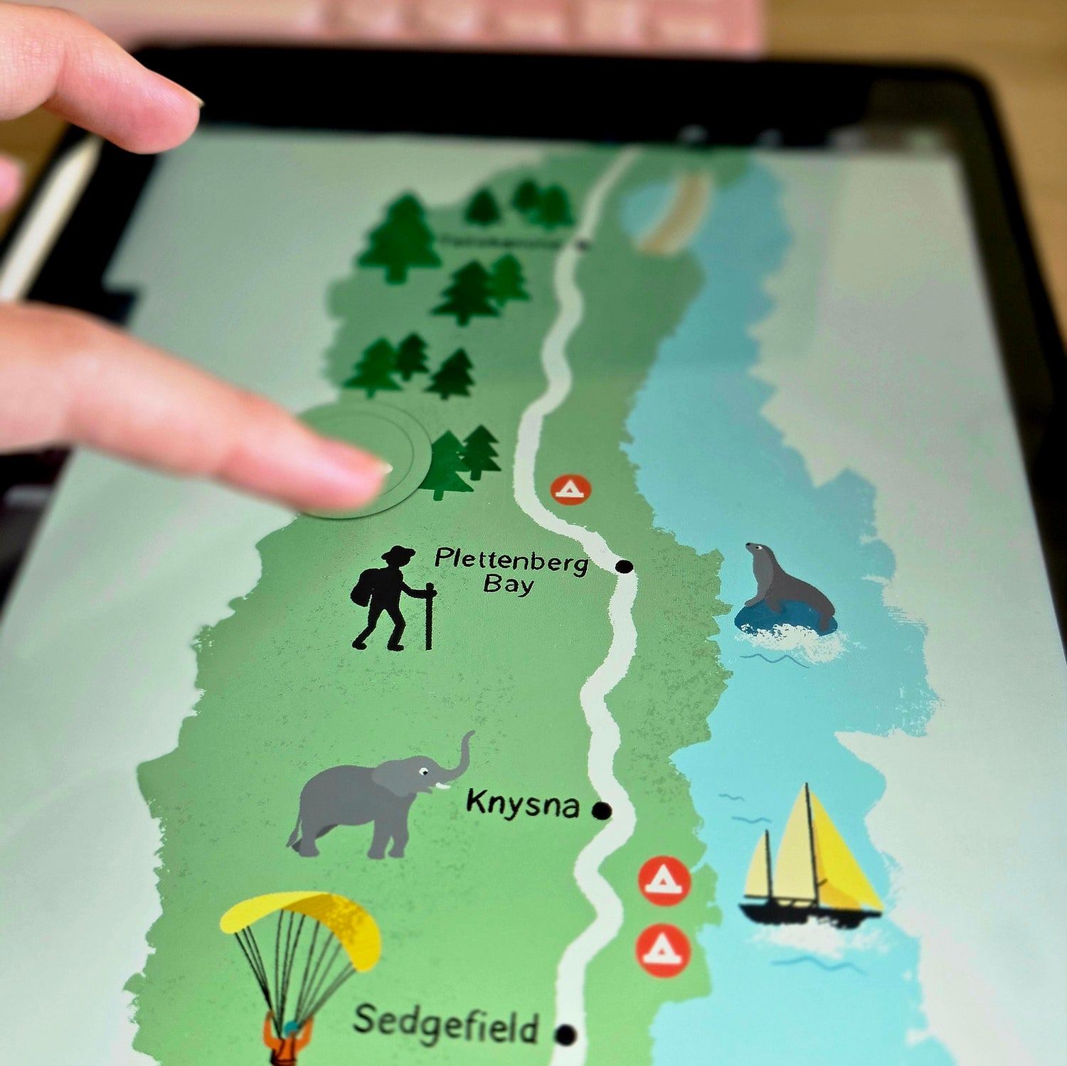 Illustrated map displayed on a ipad with Chelsea's finger pointing at it, showing locations like Plettenberg Bay, Knysna, and Sedgfield. Garden Route illustration